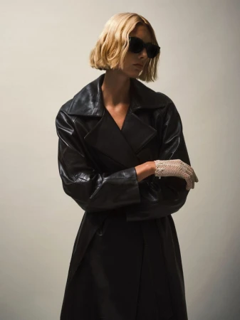 Blonde model in a black leather trench coat and vintage gloves poses against a neutral backdrop wearing dark sunglasses in a studio.