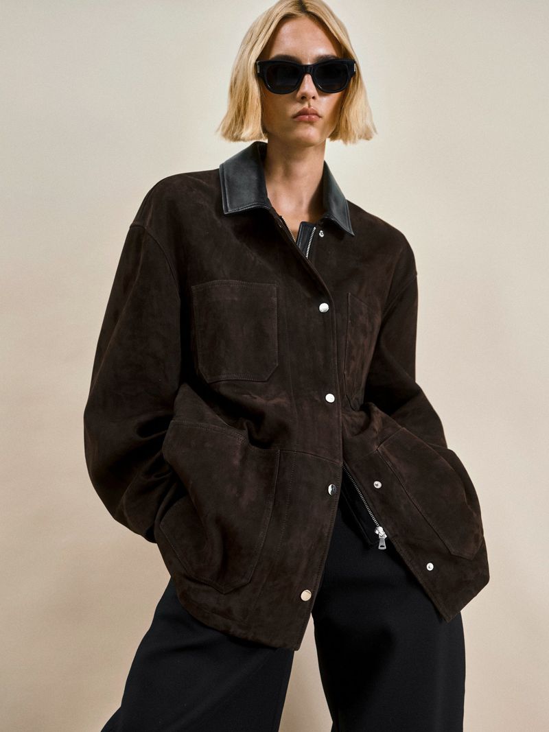 A woman wears a brown suede jacket, black top, and sunglasses against a neutral backdrop, showcasing a blend of casual and chic style.
