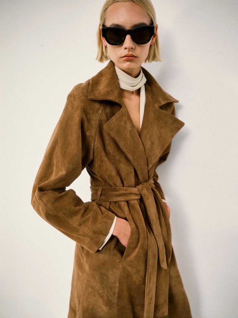 An adult woman poses in a full-length brown suede belted trench coat and black sunglasses against a plain white background in studio lighting.