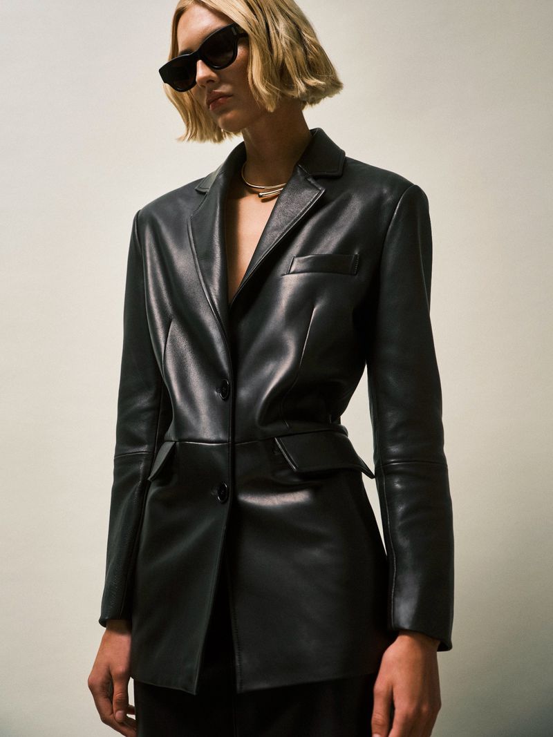 A woman in a black leather blazer and sunglasses stands against a neutral backdrop, showcasing fashion-forward style and elegant outerwear.