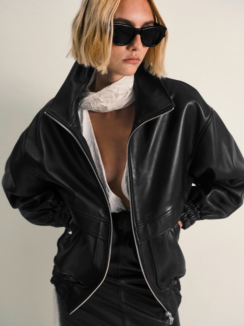 Fashionable woman with blonde bob, wearing sunglasses, leather jacket, and scarf against a neutral backdrop, looking stylish and modern.