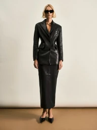 Model in black leather blazer and matching skirt wearing dark sunglasses against a seamless neutral backdrop.