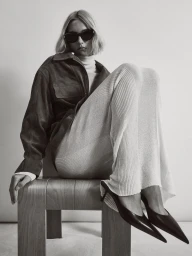 Model wearing dark sunglasses and a dark jacket seated on a wooden chair against a plain white background.