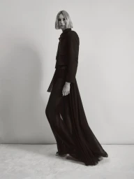 Model in black jacket and sheer pleated maxi skirt standing against a plain light background for Nour Hammour Spring Summer 25.