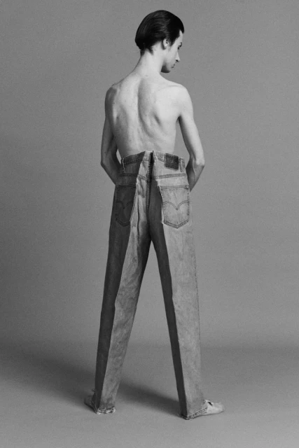 Black and white studio shot of a shirtless man standing in front of a pair of jeans with one leg in each pair, back to the camera.