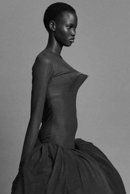 Elegant Black woman poses in long-sleeve black dress with textured tulle skirt, against a neutral gray backdrop in studio lighting.