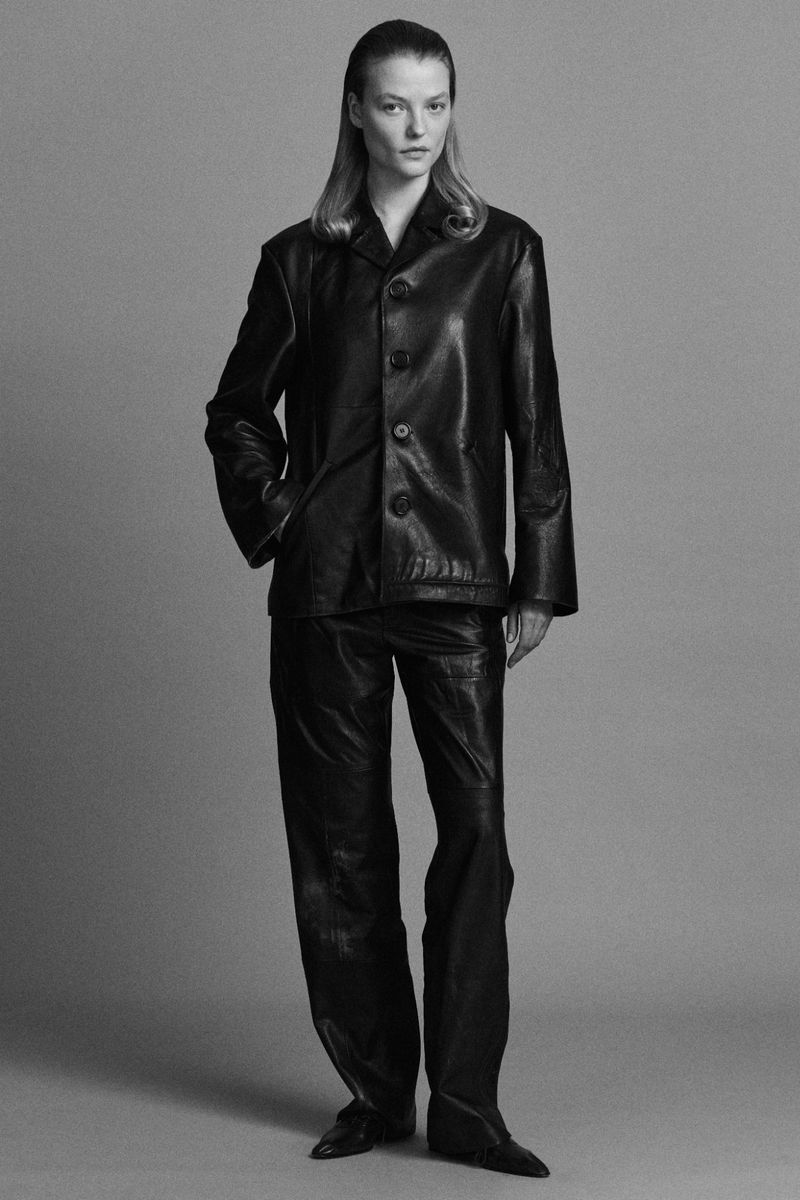Model wearing a black leather button-up jacket and matching leather trousers against a solid grey studio background.