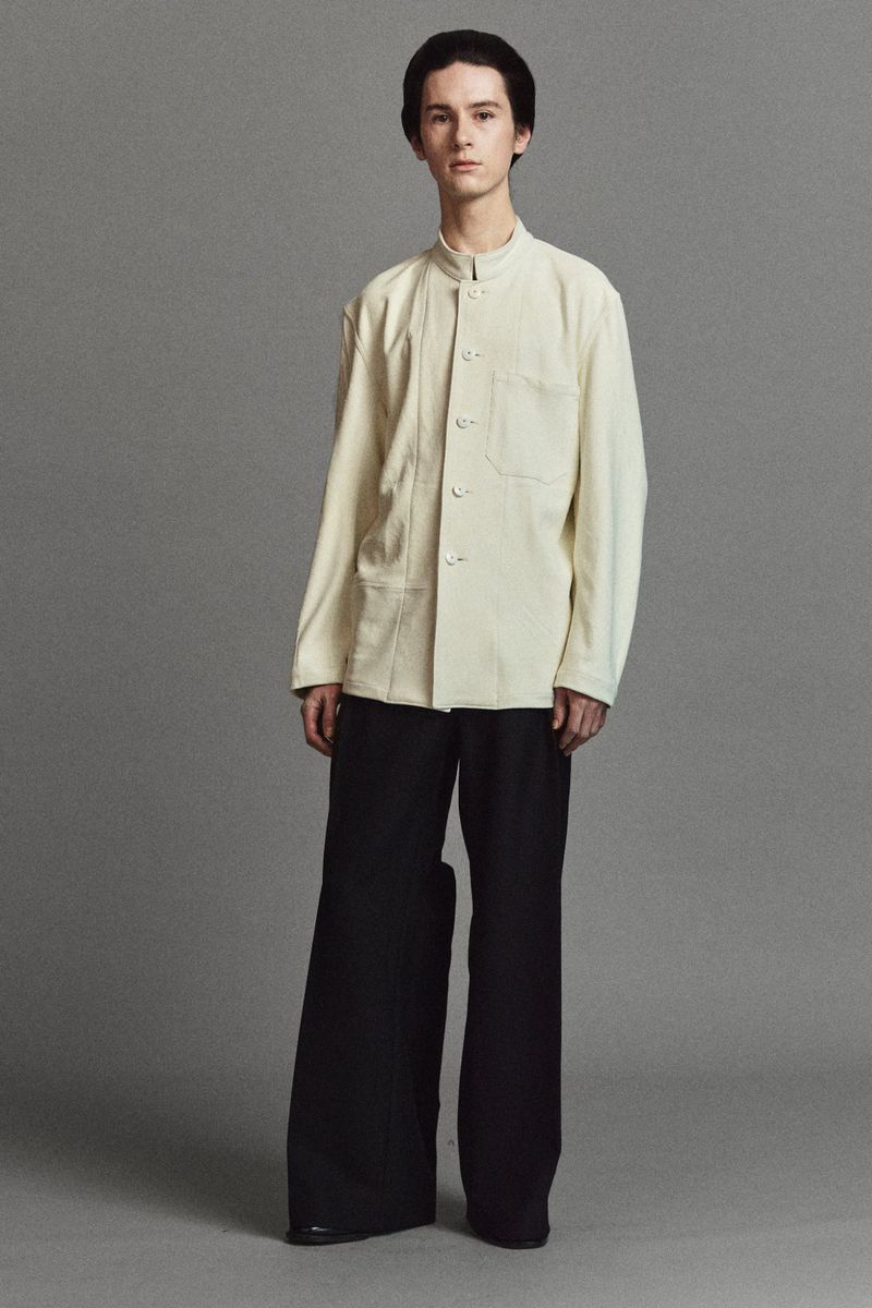 Studio shot of a young adult man in a cream-colored jacket and wide-leg black pants, posing against a neutral gray background.