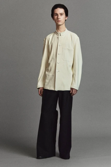 Studio shot of a young adult man in a cream-colored jacket and wide-leg black pants, posing against a neutral gray background.