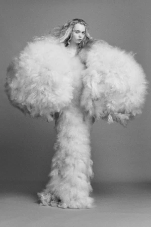 Model wearing a voluminous fluffy white textured garment against a plain grey studio backdrop
