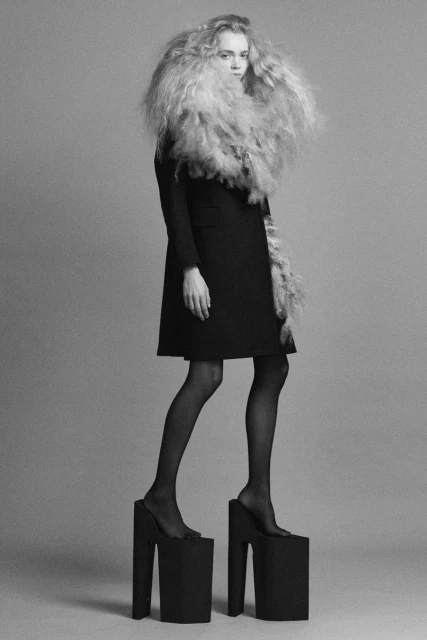 A woman in a black coat and platform shoes stands in a photo studio with a large curly hair style and feathery boa around her neck.