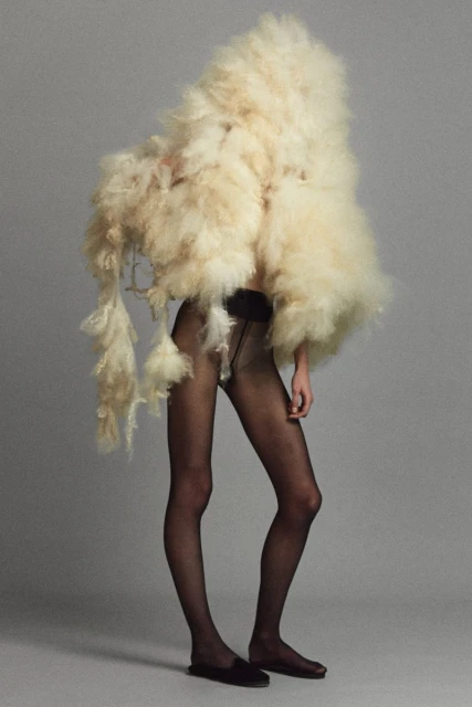 Side-shot of an adult female posing in a studio wearing black hosiery and flats, and an oversized cream-colored coat of fluffy material.