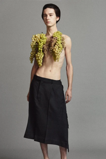 A shirtless young adult male with grapes around his neck and a black skirt poses in a studio against a grey background.