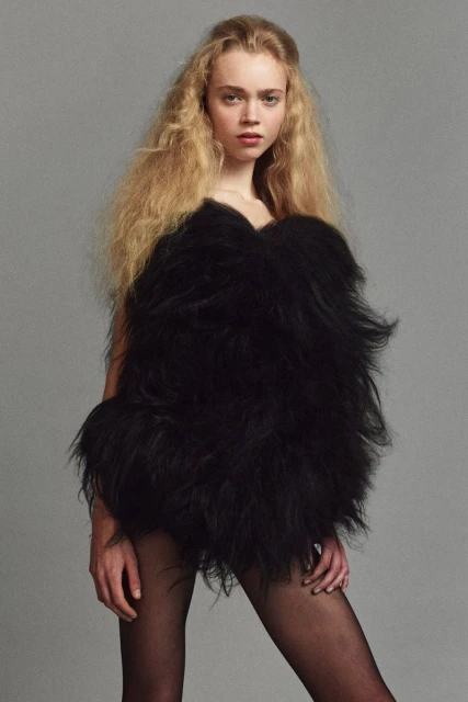 Fashion shot of a woman with long blonde hair wearing an oversize black feather boa and black tights, posing against a grey background.