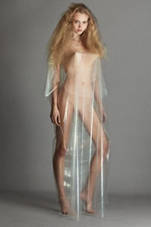 Adult female fashion model in a transparent plastic dress stands in a full shot in a studio with neutral grey backdrop.