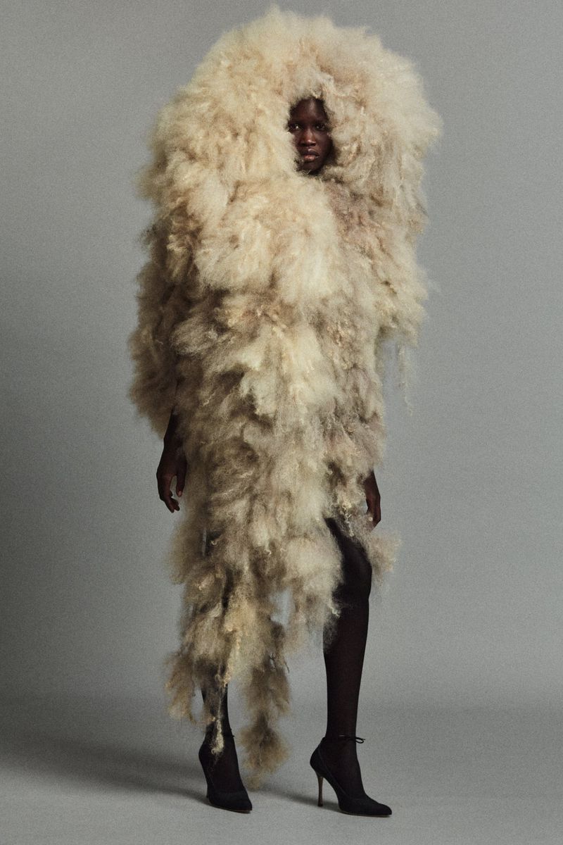 Fashion portrait of a Black model wearing an extravagant, shaggy light-beige faux-fur outfit with black stiletto heels, against a grey studio backdrop.