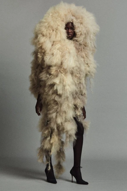 Fashion portrait of a Black model wearing an extravagant, shaggy light-beige faux-fur outfit with black stiletto heels, against a grey studio backdrop.