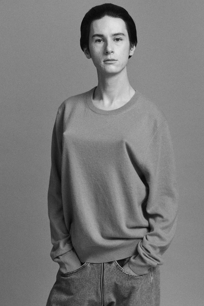 Black-and-white portrait of a slender young adult wearing a crewneck sweater and jeans, standing in a neutral indoor setting.