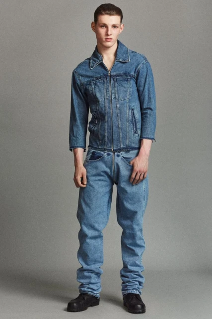 Full-length shot of a young adult man in a denim jacket and jeans, standing against a grey backdrop in a fashion editorial style.