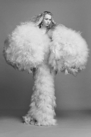 Model wearing a voluminous fluffy white textured garment against a plain grey studio backdrop