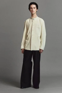 Full-length portrait of a male model wearing a cream buttoned jacket and dark trousers against a grey studio backdrop.