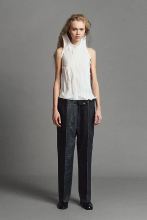 Model wearing a white structured top and dark trousers against a grey studio backdrop
