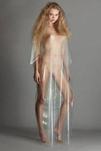 Model wearing a translucent vinyl dress and voluminous blonde hair standing against a grey studio backdrop
