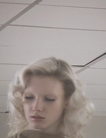 Close-up portrait of a woman with blonde wavy hair against a plain ceiling panel background