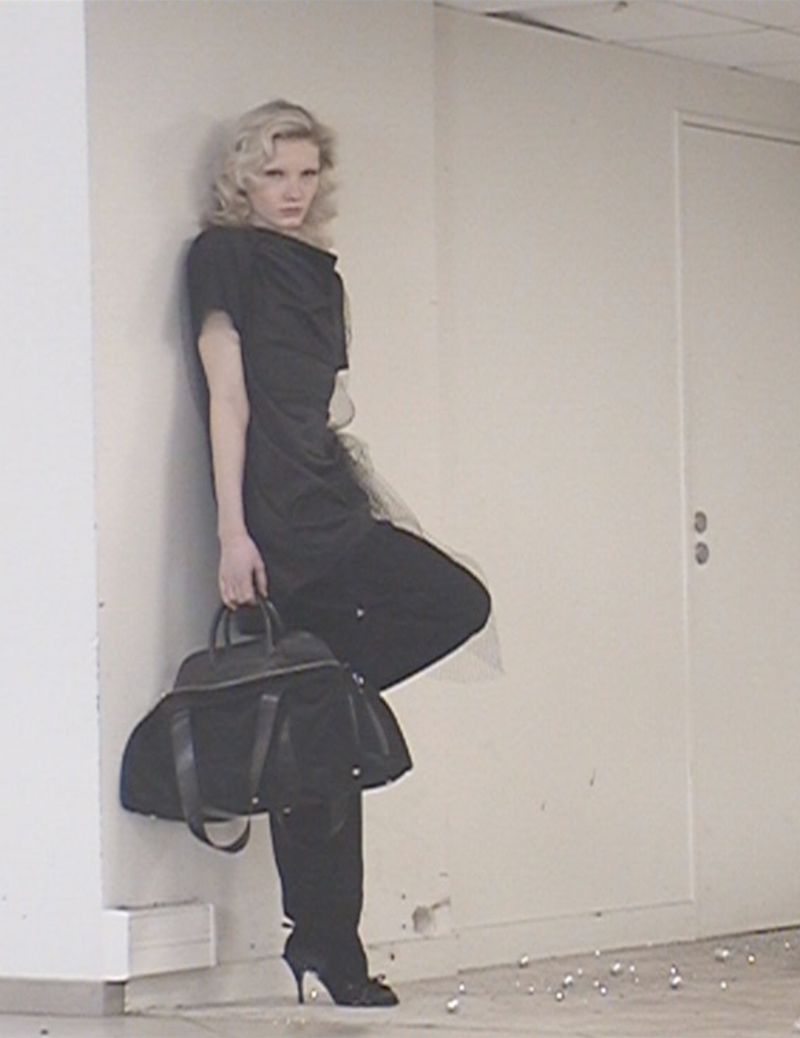 Blonde woman in dark dress leans against wall holding black handbag in an indoor space with a door and small, round decorative objects.