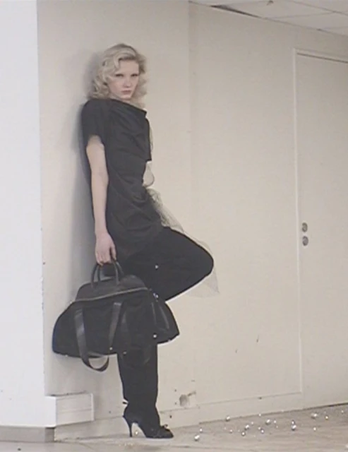Blonde woman in dark dress leans against wall holding black handbag in an indoor space with a door and small, round decorative objects.