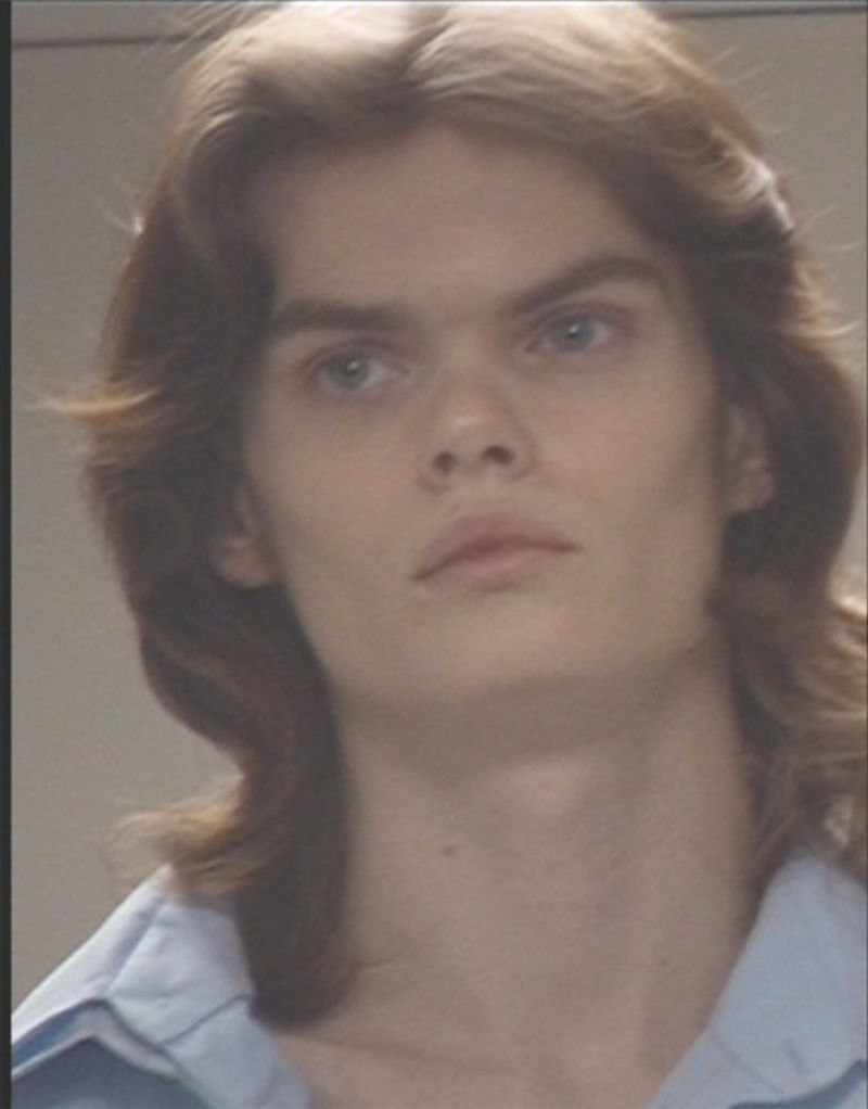 Portrait of a young adult with long brown hair, wearing a light blue collared shirt, against a plain, soft-focus background.