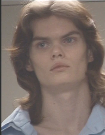 Portrait of a young adult with long brown hair, wearing a light blue collared shirt, against a plain, soft-focus background.