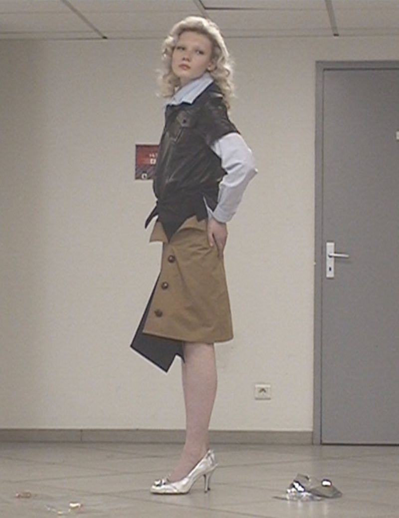 Fashionable young adult woman poses in modern outfit with blonde wavy hair, wearing a short black jacket and beige buttoned skirt.
