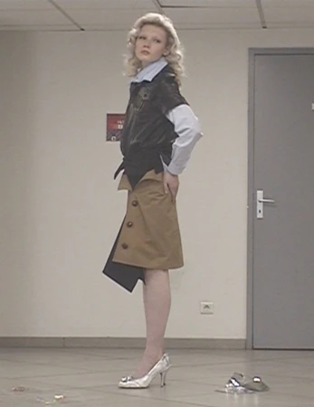 Fashionable young adult woman poses in modern outfit with blonde wavy hair, wearing a short black jacket and beige buttoned skirt.