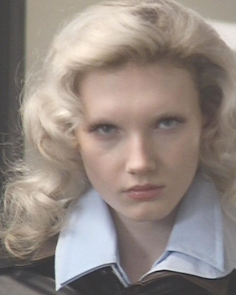Close-up portrait of a fair-skinned young woman with blonde curly hair, wearing a collared shirt and jacket in a neutral indoor setting.