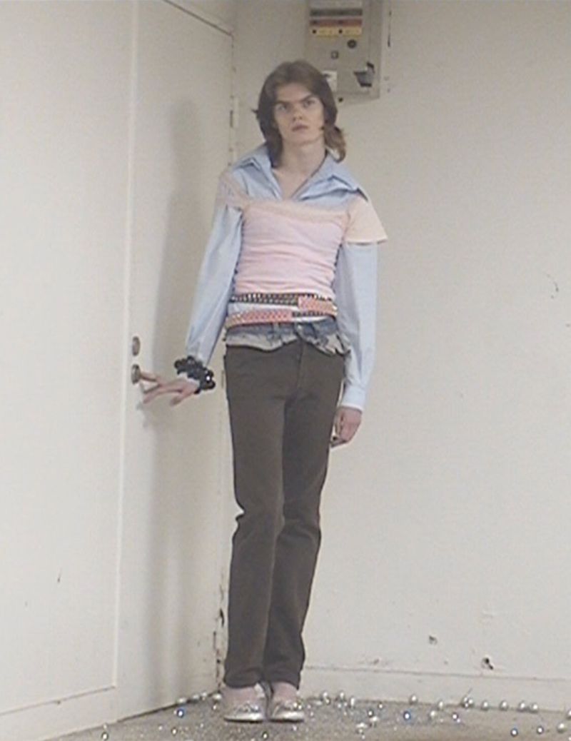 Full-length portrait of a person with long brown hair in an eclectic outfit, standing indoors with a hand near a door.