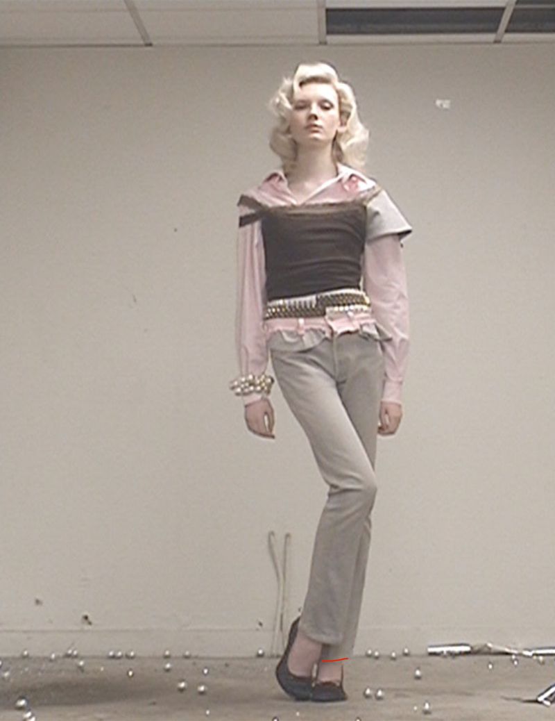 A fair-skinned fashion model poses in a studio with soft lighting, wearing a brown bustier, pink top, jeans, ballet flats, and vintage jewelry.