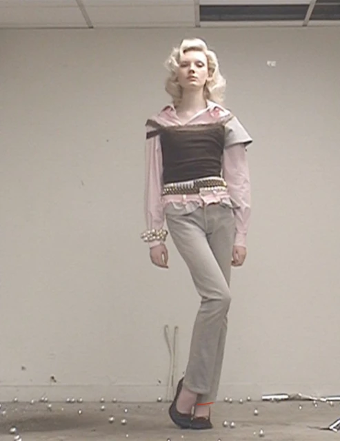 A fair-skinned fashion model poses in a studio with soft lighting, wearing a brown bustier, pink top, jeans, ballet flats, and vintage jewelry.