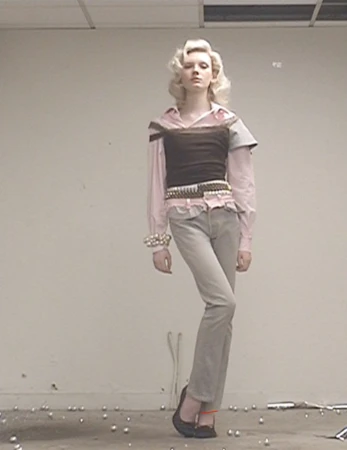 A fair-skinned fashion model poses in a studio with soft lighting, wearing a brown bustier, pink top, jeans, ballet flats, and vintage jewelry.