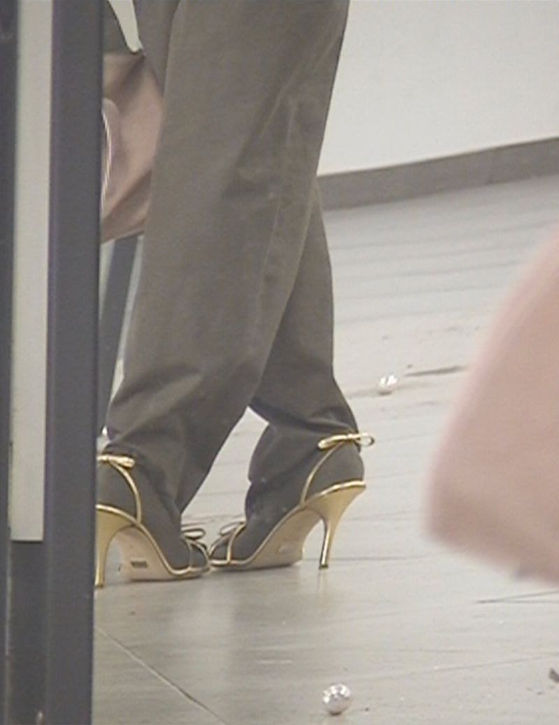 View of an adult wearing brown trousers and gray high-heeled sandals, with a gold heel. Some pearls are scattered on the floor.