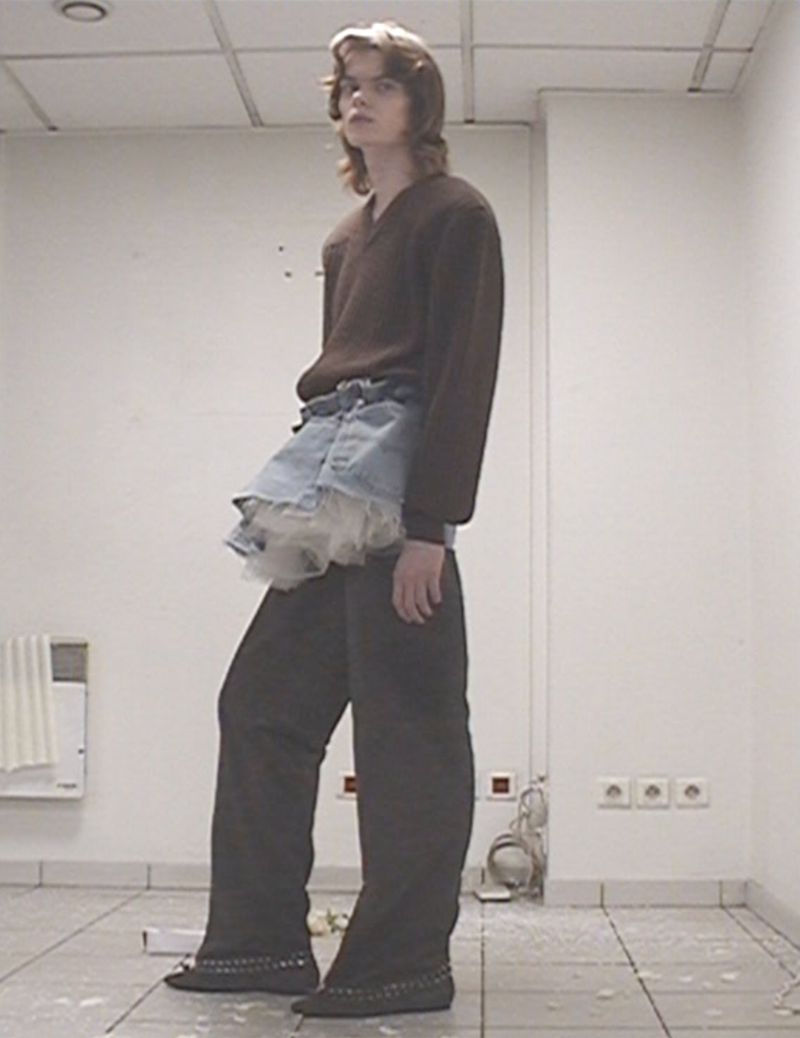 Full-length shot of an adult with long hair wearing a brown sweater, denim skirt with lace, and dark pants, standing in a white room.