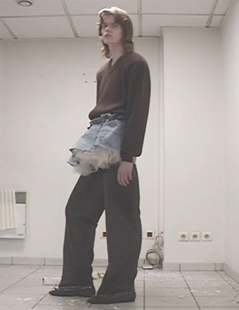 Full-length shot of an adult with long hair wearing a brown sweater, denim skirt with lace, and dark pants, standing in a white room.