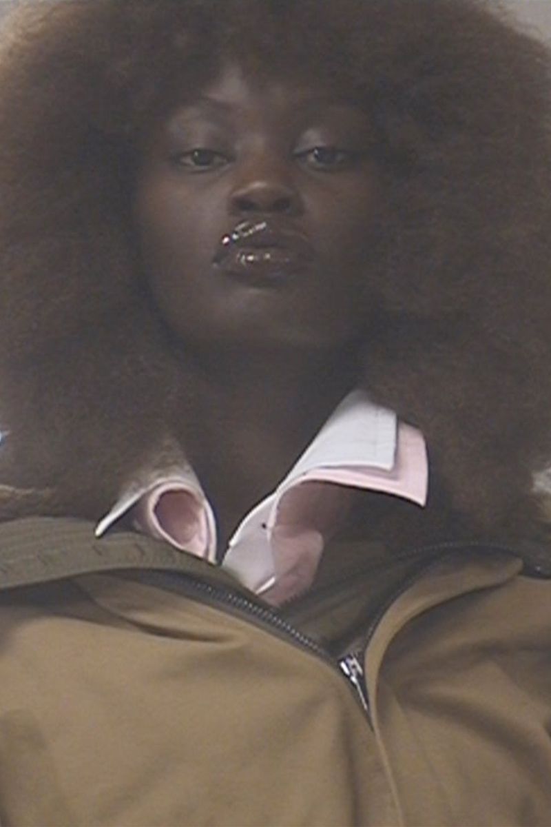 Close-up portrait of an African woman with afro hairstyle wearing a collared shirt and tan jacket, captured indoors with natural lighting.