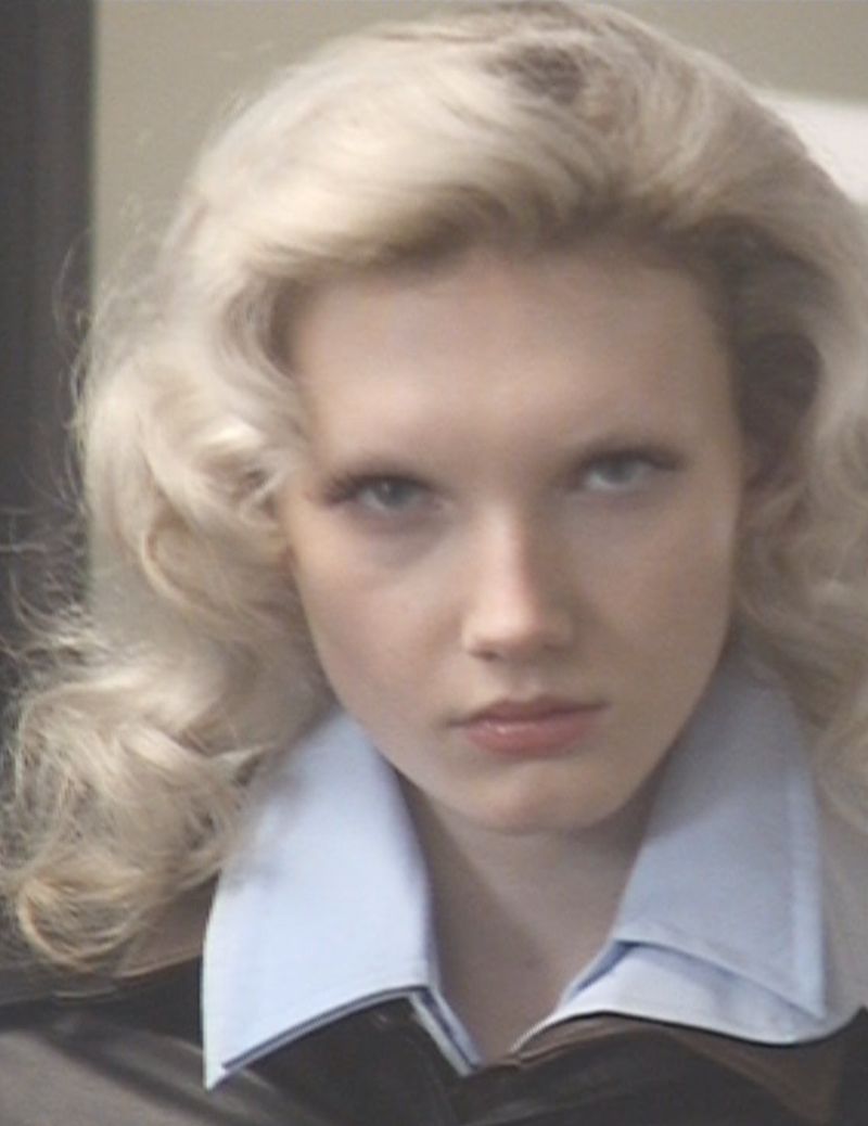 Close-up portrait of a blonde model with voluminous hair wearing a light blue collared shirt and dark jacket.