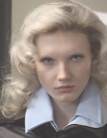 Close-up portrait of a blonde model with voluminous hair wearing a light blue collared shirt and dark jacket.