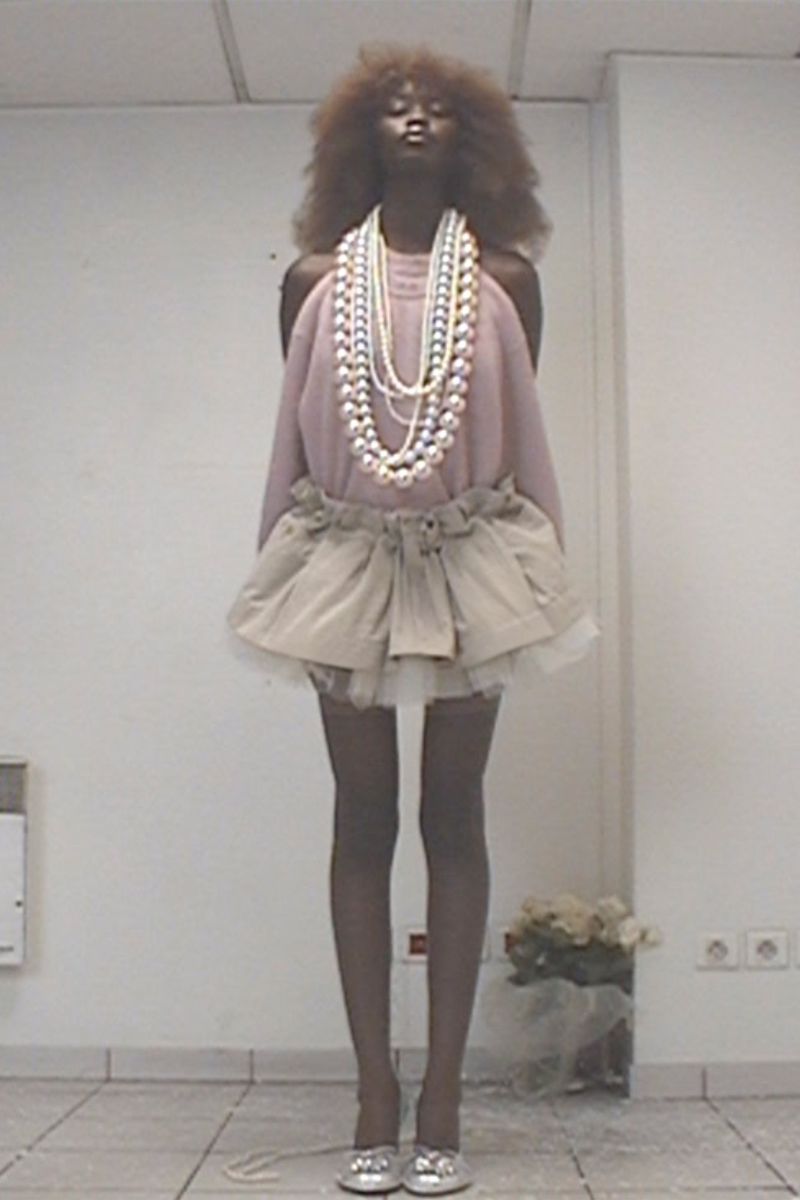 Elegant woman with afro, pearls, tutu, and silver flats poses in a stark white room. High fashion meets theatrical whimsy in a contemporary setting.