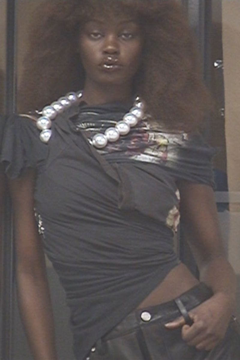 Fashion portrait of a woman with curly brown hair wearing a pearl necklace, draped black top, and leather pants, posing in an indoor setting.