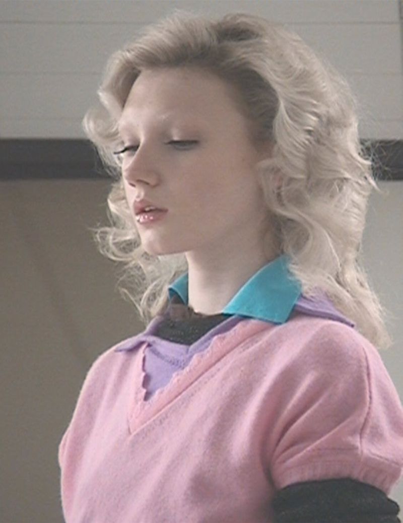 Portrait of fair-skinned blonde woman with eyes closed wearing a pink sweater with blue collar, against a neutral interior background.