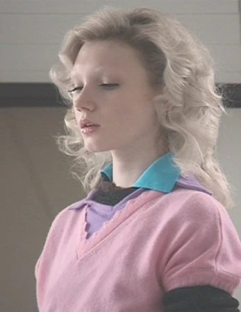 Portrait of fair-skinned blonde woman with eyes closed wearing a pink sweater with blue collar, against a neutral interior background.