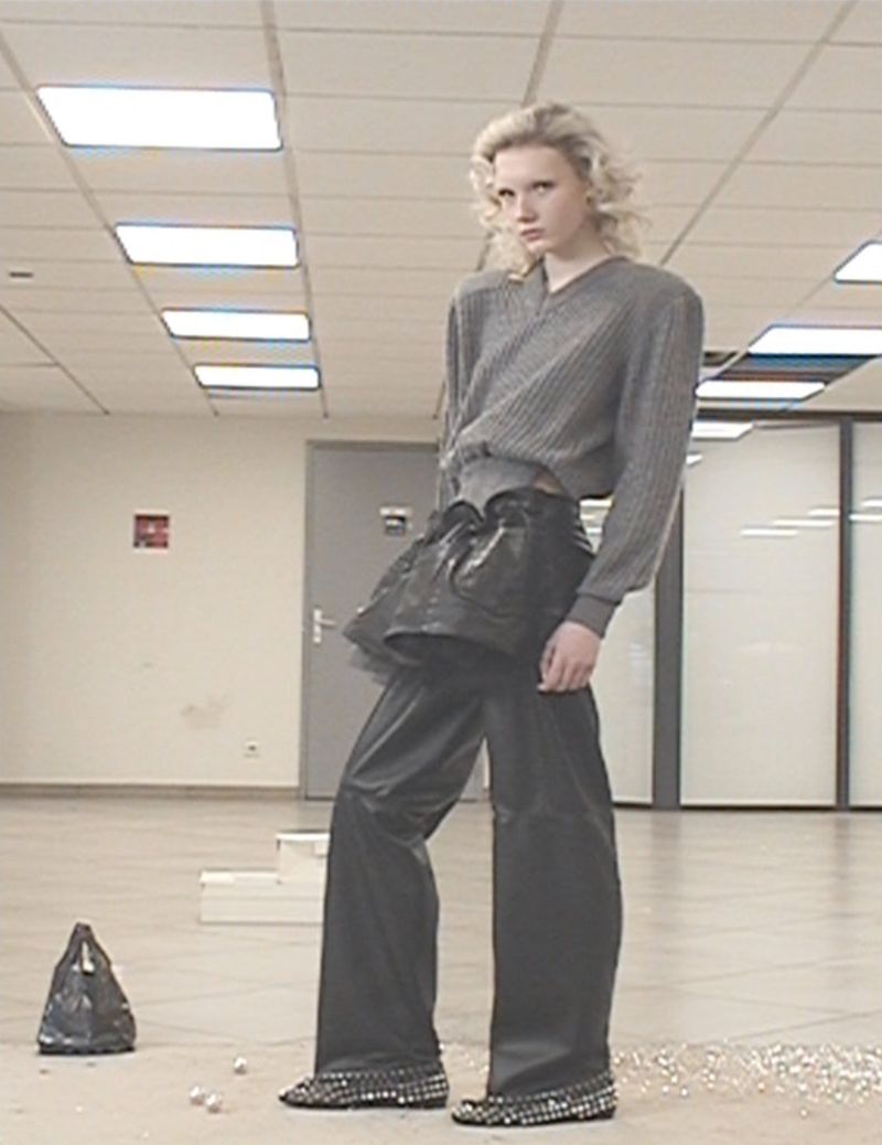 Fashion model poses indoors, wearing grey sweater, sculptural black skirt, wide black trousers, and embellished shoes. A black bag sits nearby.